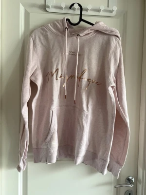 Ljusrosa hoodie med texttryck - Ljusrosa hoodie med huva och dragsnören, samt magficka framtill. På bröstet står det 'C'est SIMPLEMENT Magnifique' i guldigt tryck. Tillverkad i mjukt bomullsmaterial och har ribbade muddar vid ärmslut och nederkant. Perfekt för en chill och avslappnad stil.
