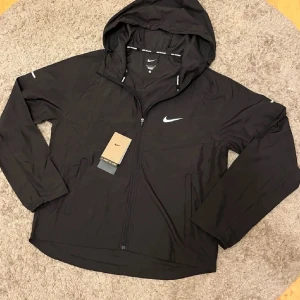Nike Miller - Snygg svart vindjacka från Nike med huva och dragkedja framtill. Jackan har vita Nike-loggor på bröstet och ärmen samt justerbar huva. Perfekt för blåsiga dagar och sportiga outfits.