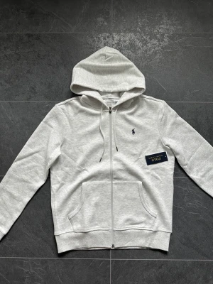 Ljusgrå hoodie från Polo Ralph Lauren - Snygg ljusgrå hoodie från Polo Ralph Lauren med dragkedja, huva med dragsko och klassisk logga broderad på bröstet. Tillverkad i mjuk bomullsblandning och har två stora fickor framtill. Perfekt för en avslappnad och stilren look.