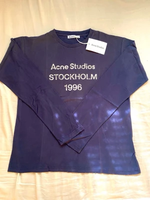 Blå långärmad topp Acne Studios - Mörkblå långärmad topp från Acne Studios med tryck 'Acne Studios STOCKHOLM 1996' på bröstet. Klassisk rund halsringning och relaxed fit. Perfekt för dig som gillar minimalistisk och stilren design.