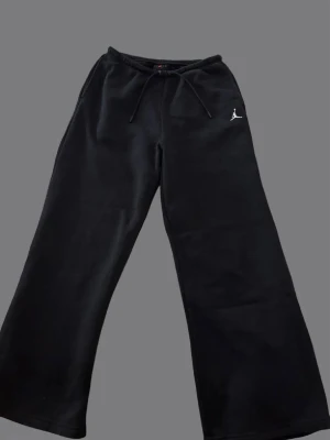 Svarta Jordan mjukisbyxor med brodyr - Säljer ett par svarta mjukisbyxor från Jordan med breda ben och dragsko i midjan. Byxorna har en vit broderad Jumpman-logga på vänster sida och är gjorda i mjukt, bekvämt material. Perfekta för chill dagar eller streetwear-stil.