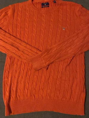 Orange kabelstickad tröja från Gant - Snygg orange kabelstickad tröja från Gant i premium bomull. Tröjan har rund halsringning, ribbade muddar och Gant-logga broderad på bröstet. Perfekt för dig som vill ha en färgstark och klassisk look.