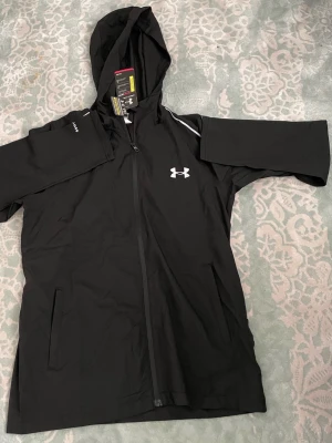 Svart vindjacka från Under Armour - Snygg svart vindjacka från Under Armour med huva och hel dragkedja framtill. Jackan har vita detaljer vid axlarna och Under Armour-logga på bröstet. Tillverkad i lätt och funktionellt material, perfekt för blåsiga dagar. Två fickor framtill och justerbar huva.