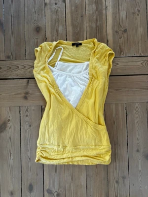 Gul omlottopp från Vanilla - Snygg gul omlottopp från Vanilla med vit insydd topp under. Kortärmad modell med avslappnad passform och omlottdesign framtill. Perfekt för dig som gillar färg och vill ha en unik detalj i garderoben.