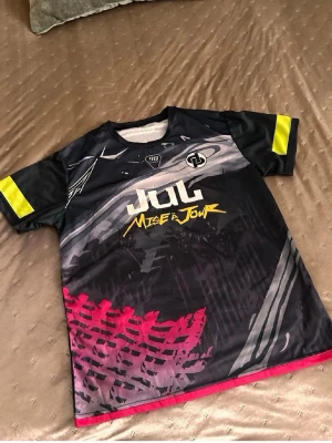  t-shirt från JUL - Cool svart t-shirt från JUL med grafiskt tryck i vitt, gult och rött samt texten 'Miser Jour' på framsidan. T-shirten har korta ärmar med gula detaljer och är tillverkad i ett lätt och skönt polyestermaterial. Perfekt för dig som gillar streetwear och unika prints.