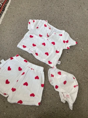 Vitt set med röda hjärtan från SHEIN - Sött tre-delat set från SHEIN i vitt med röda hjärtan. Setet består av en kortärmad topp med knappar, shorts med resår i midjan och en matchande scarf. Tillverkat i mjukt, tunt bomullsliknande tyg som känns luftigt och bekvämt.