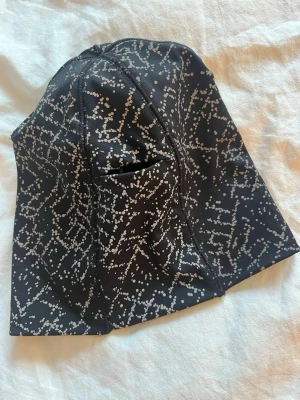 Svart mössa med reflex mönster - Snygg svart mössa med vitt prickigt och linjärt mönster som ger en modern känsla. Tillverkad i stretchigt material som sitter skönt på huvudet. Perfekt accessoar för våren. El till löpspåret el när du kör längd .  