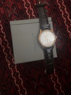 GANT klocka med brunt läderband - Stilren klocka från GANT med vitt urtavla, roséguldfärgat boett och brunt läderband med krokodilpräglat mönster. Klassisk design med minimalistiska index och separat sekundvisare. Perfekt accessoar för dig som gillar tidlös stil.