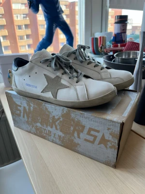 Vita Golden Goose sneakers med stjärna - Säljer ett par vita Golden Goose sneakers med grå mockastjärna på sidan, grå skosnören och svart detalj på hälen. Skorna har en klassisk låg siluett och är tillverkade i läder med mockadetaljer.     Pris kan alltid diskuteras😁