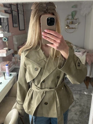Le Magda Kort beige trenchcoat med knytskärp - Stilren kort trenchcoat i beige med dubbelknäppning och brett knytskärp i midjan. Klassisk krage och axelklaffar ger en trendig look. Jackan har långa ärmar med slejfdetaljer vid ärmslut. Perfekt för dig som gillar minimalistisk och tidlös stil.