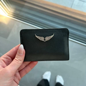 Zadig & Voltaire korthållare - Snygg svart korthållare i läder från Zadig & Voltaire med silverfärgad strass vingdetalj på framsidan. Flera fack för kort på baksidan. Försiktigt använd.