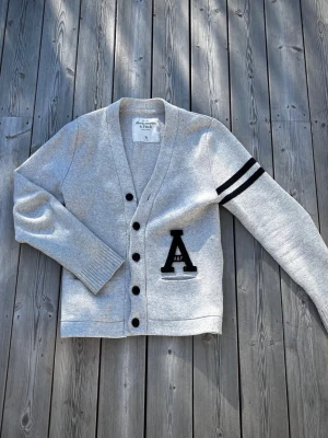 Abercrombie & Fitch cardigan - En riktigt fet grå och svart cardigan från märket Abercrombie & Fitch. Den är stickad i ett skönt och snyggt material med svarta detaljer som ränderna på ärmen och logan. Den har inga defekter och är allmänt fräsch. Storleken är Xl men jag skulle rekommendera den om du har L eftersom måtten är längd: 67cm och bredd:47cm. Kom pm för frågor eller annat.