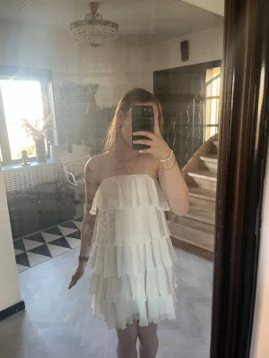 Vit axelbands volangklänning - Säljer en superfin vit kortklänning med flera lager volanger och axelbandslös design. Klänningen har en luftig och flowy känsla, perfekt för varma dagar. Materialet känns lätt och svalt, och modellen ger en romantisk vibe. Passar s-xs