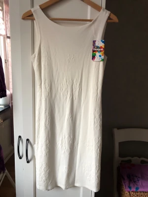 Stilren Vit kortklänning från Desigual - Säljer en vit kortklänning från Desigual med subtila broderade blommönster. Klänningen är ärmlös och har en rundad halsringning både fram och bak. Materialet känns mjukt och stretchigt, perfekt för dig som gillar stilrena och enkla plagg.