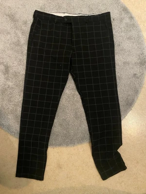 Svarta kostymbyxor med rutmönster - Snygga svarta kostymbyxor med vitt rutmönster och klassisk passform. Byxorna har hällor för bälte, knapp och dragkedja framtill samt sidfickor. Perfekta för en stilren look.