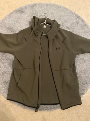 Olivgrön hoodie från Under Armour - Snygg olivgrön hoodie från Under Armour med dragkedja framtill och svart logga på bröstet. Hoodien har huva, två sidofickor och svarta detaljer vid axlarna. Tillverkad i ett mjukt och stretchigt material som passar perfekt till vardags eller träning.