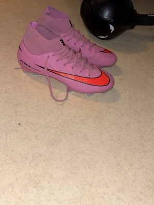 Rosa Nike Mercurial fotbollsskor - Säljer ett par rosa Nike Mercurial fotbollsskor med högt skaft och snörning. Skorna har en svart och röd swoosh-logga på sidan samt detaljer i svart. Yttermaterialet är syntet och sulan har fasta dobbar för spel på gräs.
