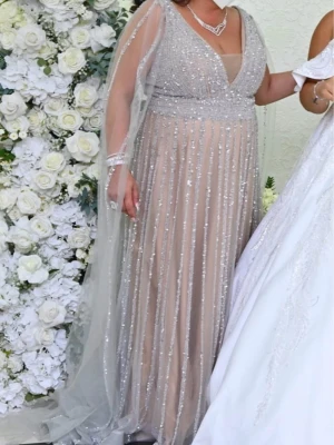 Cat walk Glittrig långklänning med släp - Elegant långklänning i beige och silver med glittriga paljetter och transparenta ärmar. Djup v-ringning och markerad midja, samt ett skirt släp som ger extra glamour. Perfekt för speciella tillfällen.