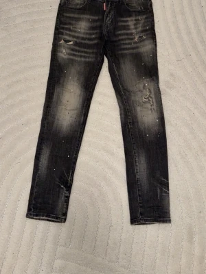 Dsquared jeans  - Säljer ett par svarta skinny jeans med slitningar och distressed detaljer på både fram- och baksida. Jeansen har en tvättad look med ljusare partier och små färgstänk för en trendig, avslappnad stil. Perfekta för dig som gillar streetwear och edgy mode.