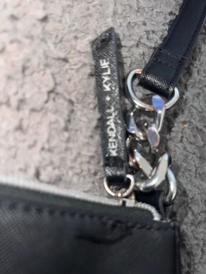 Svart handväska Kendall + Kylie - Snygg svart handväska från Kendall + Kylie med stilren rektangulär form. Väskan har en axelrem med silverfärgade kedjedetaljer och är gjord i ett tåligt konstlädermaterial. Perfekt accessoar för att lyfta din outfit.