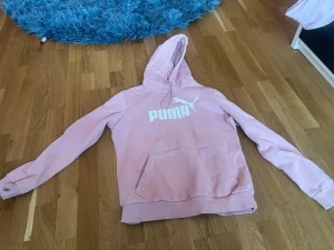 Rosa hoodie från Puma med huva - Säljer en ljusrosa hoodie från Puma med stor vit logga på bröstet. Hoodien har huva, magficka och ribbade muddar vid ärmslut och nederkant. Perfekt för en avslappnad och sportig stil.