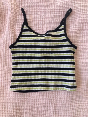 Randig ribbad linnetopp - Säljer en croppad linnetopp med smala axelband och ribbad struktur. Toppen har svarta, vita och ljusgula horisontella ränder och är i ett mjukt stretchigt material. Perfekt till sommaren och enkel att matcha med jeans eller kjol.