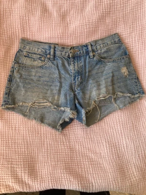 Blå jeansshorts med fransig kant - Säljer ett par blå jeansshorts från 157 med fransig kant och slitna detaljer framtill. Shortsen har klassisk femficksdesign, knapp och dragkedja i midjan samt bakfickor. Perfekta för varma dagar och har en avslappnad loose passform.