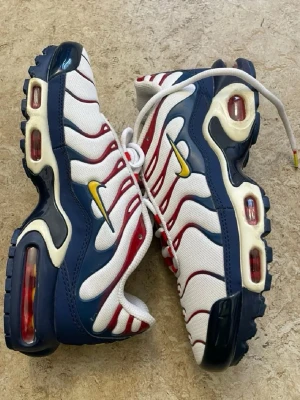 Nike Air Max Plus TN blå/vit/röd - Nike Air Max Plus TN sneakers i blått, vitt och rött med gula detaljer. Ovandelen är i mesh med vågiga röda linjer och den ikoniska gula Swoosh-loggan. Synliga Air-enheter i sulan och robust yttersula för extra komfort och stil.