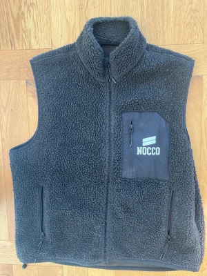 Svart fleeceväst från NOCCO - Mysig svart fleeceväst från NOCCO med hög krage och hel dragkedja. Västen har två sidofickor med dragkedja och en bröstficka med NOCCO-logga och dragkedja. Perfekt för lager-på-lager och chill stil. Passar L/XL