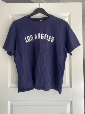Svart t-shirt med Los Angeles-tryck - Svart t-shirt med vit text 'LOS ANGELES' tryckt på bröstet. Klassisk rund hals och korta ärmar. T-shirten är i mjuk bomull och har en avslappnad passform som passar perfekt till jeans eller shorts.