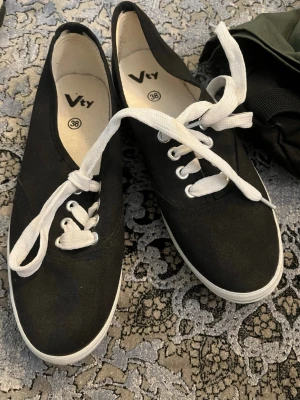 Svarta låga sneakers  - Klassiska svarta sneakers med vita snören och vit sula. Skorna har en enkel och stilren design med lågt skaft och textilmaterial. Perfekta för en avslappnad och trendig look.