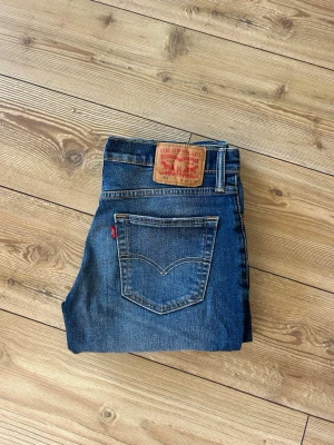 Levi's blå raka jeans - Klassiska blå jeans från Levi's med rak passform och fem fickor. Jeansen har en tydlig läderpatch bak i midjan och den ikoniska röda Levi's-taggen på bakfickan. Tillverkade i slitstark denim med snygg tvätt och subtila slitningar.