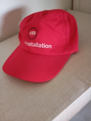 Röd keps från ABB Installation - Snygg röd keps med broderad ABB-logga och texten 'Installation' framtill. Klassisk böjd skärm och justerbar passform bak. Tillverkad i bomullstyg som andas. Perfekt för dig som gillar industriell stil eller vill sticka ut med en unik accessoar.