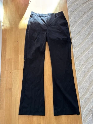 Svarta kostymbyxor från Lindex - Snygga svarta kostymbyxor från Lindex med klassisk rak passform och diskret knappstängning i midjan. Byxorna har bälteshällor och är tillverkade i ett mjukt tyg som ger en stilren look. Perfekta för dig som vill ha en clean och enkel stil. Modell ”Fiona”