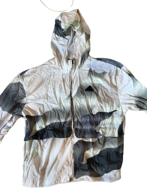 Adidas vindjacka med camomönster - Snygg vindjacka från Adidas med huva och dragkedja framtill. Jackan har ett unikt camomönster i vitt, grått och svart samt Adidas-logga på bröstet. Tillverkad i lätt och tunt material som passar perfekt för blåsiga dagar.