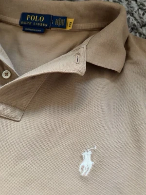 Beige pikétröja från Polo Ralph Lauren - Klassisk beige pikétröja från Polo Ralph Lauren med vit broderad logga på bröstet. Tröjan har krage, knappar vid halsen och är tillverkad i bomull för en bekväm känsla. Perfekt till jeans eller shorts för en clean look.🚨priset kan alltid diskuteras🚨skriv om ni har frågor!