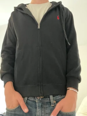Svart hoodie från Polo Ralph Lauren - Svart zip hoodie från Polo Ralph Lauren med röd logga broderad på bröstet. Tröjan har huva med snörning, dragkedja framtill och två fickor. Det är storlek M men den har krympt i tvätten så den passar mer XS/S än M. Hör av er vid funderingar!