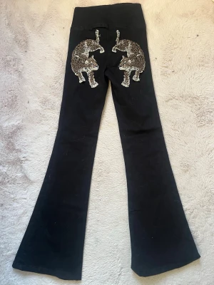 Svarta jeans med leopardbrodyr - Unika svarta jeans med utsvängda ben och två stora leopardbroderier på bakfickorna. Snygg detalj som sticker ut och ger en cool vibe till din outfit. Perfekta för dig som vill ha något annorlunda och trendigt.