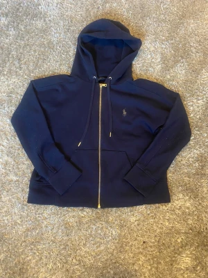 Mörkblå hoodie från Polo Ralph Lauren - Snygg mörkblå hoodie från Polo Ralph Lauren med dragkedja framtill, justerbar huva och klassisk logga broderad på bröstet. Tillverkad i mjukt bomullsmaterial och har fickor framtill. Perfekt för en avslappnad och stilren look.