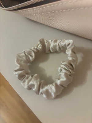 Ljusbeige scrunchie i satin - Säljer en snygg ljusbeige scrunchie i glansigt satinmaterial. Perfekt för att ge en extra touch till din frisyr och skonsam mot håret tack vare det mjuka tyget. Passar både till hästsvans och knut.