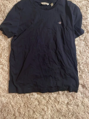 Marinblå t-shirt från GANT - Klassisk marinblå t-shirt från GANT med broderad logga på bröstet. Tillverkad i mjuk bomull och har en normal passform med korta ärmar och rund hals. Perfekt basplagg för en clean och stilren look. Jag kommer stryka den innan säljning! Endast använd 1 gång!