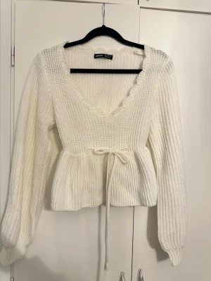 Vit stickad topp med knytband - Säljer en söt vit stickad topp med djup v-ringning, volangkant och knytband i midjan. Tröjan har långa puffärmar och ribbstickad struktur som ger en trendig och feminin look.
