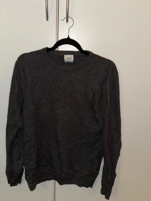 Mörkgrå stickad tröja från John Henric  - En enkel och stilren mörkgrå stickad tröja från John Henric. Tröjan har rund halsringning, långa ärmar och ribbade muddar vid ärmslut och nederkant. Perfekt basplagg i mjukt material som passar till det mesta.