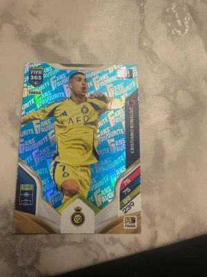 Panini FIFA 365 Adrenalyn XL kort - Säljer ett Panini FIFA 365 Adrenalyn XL samlarkort med holografisk yta. Kortet visar en fotbollsspelare i gul matchtröja och har texten 'Fans Favourite'. Baksidan har en aktiveringskod för online-spel. Perfekt för dig som samlar på fotbollskort.