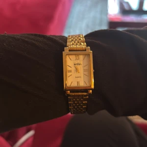 Guldfärgad rektangulär Sittar-klocka - Stilren guldfärgad klocka från Sittar med rektangulärt ur och klassisk länkarmband i metall. Urtavlan är vit med romerska siffror och tunna visare. Perfekt accessoar för dig som gillar tidlös och elegant design.