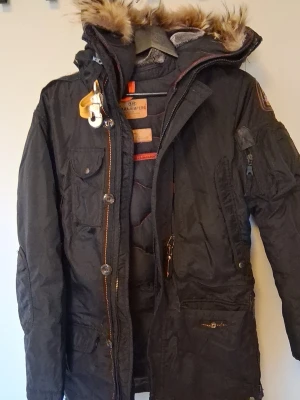 Svart parkas från Parajumpers - Säljer en svart parkas från Parajumpers med avtagbar fuskpäls på huvan, flera fickor och detaljer i orange. Jackan har dragkedja och knappar framtill samt patch på ärmen. Perfekt för kalla dagar och riktigt snygg streetstil.
