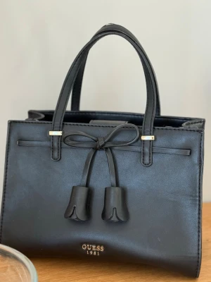 Svart handväska  - Stilren svart handväska med dekorativ rosett och tofsar framtill. Väskan har guldfärgade detaljer och logotypen 'Guess 1981' i guld längst ner. Tillverkad i slätt konstläder med en fyrkantig form. Gott om utrymme! Finns ett tillhörande band till så att man kan ha över axeln🥰något sliten, konstlädret börjar gå upp lite. FEJK Guess, ej äkta 