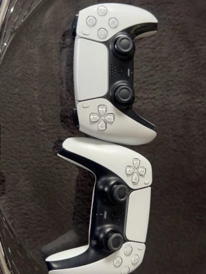 PlayStation 5 DualSense handkontroller - Två vita och svarta PlayStation 5 DualSense handkontroller med ergonomisk form, klassiska PlayStation-knappar och dubbla analoga spakar. Tillverkade i plast med detaljer i svart och vitt. Perfekt för gaming på PS5.