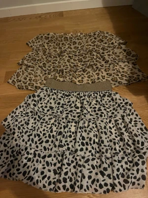 Två volangkjolar med djurmönster - Säljer två snygga volangkjolar, en med leopardmönster i beige och brunt och en med svartvitt leopardliknande mönster och glittrig midjeresår. Perfekta för att lyfta din outfit med trendiga detaljer och coola prints.