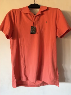 Morris  pikétröja  - Snygg korallfärgad pikétröja med klassisk krage och korta ärmar. Tröjan har en liten broderad logga på bröstet och är tillverkad i mjuk bomull som känns skön mot huden. Perfekt till jeans eller shorts för en avslappnad look.